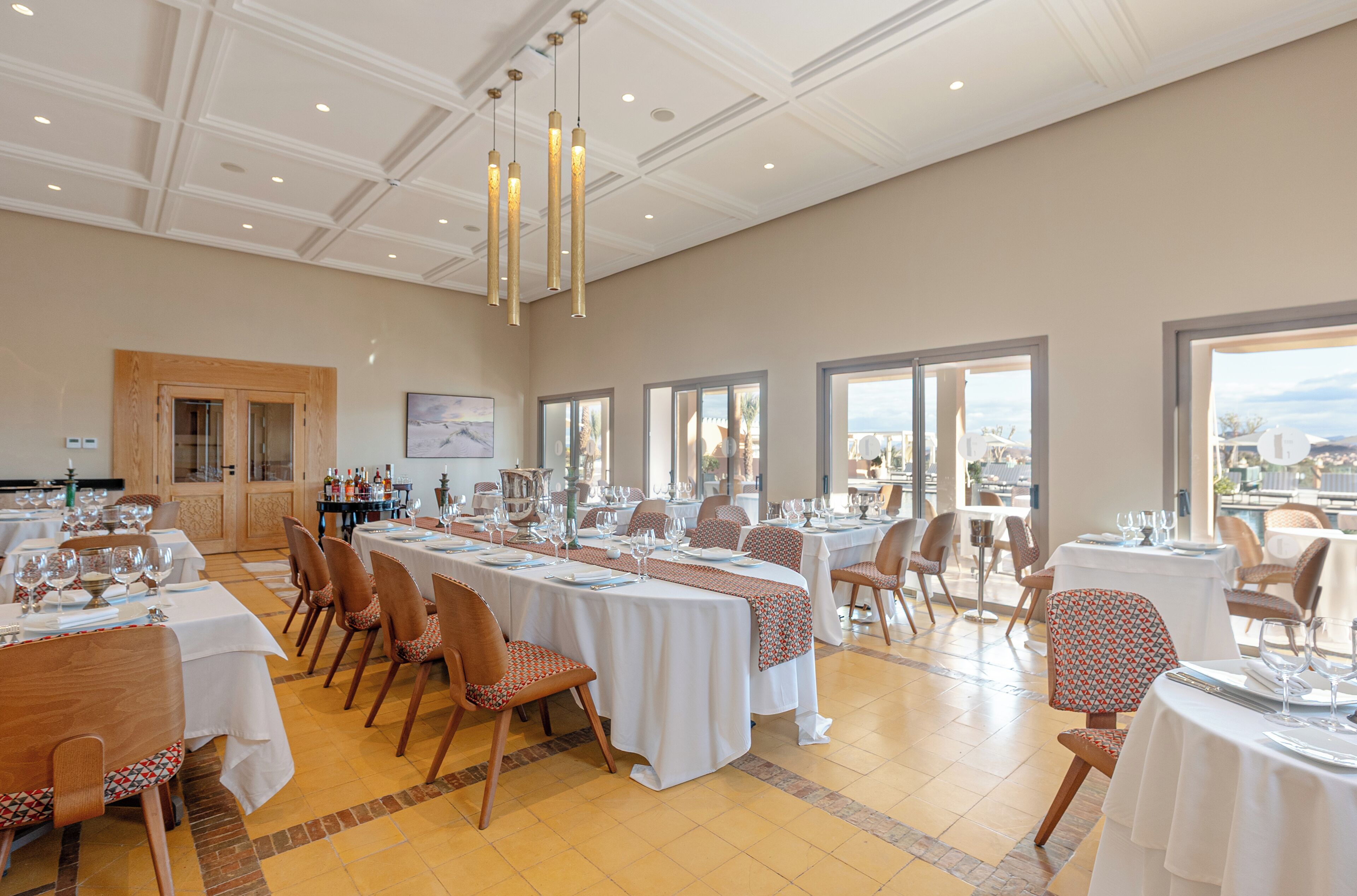 Un vaste espace dédié à la salle à manger, avec de nombreuses tables dressées pour les convives, de grandes fenêtres offrant une vue dégagée et un plafond haut agrémenté d’un éclairage décoratif.