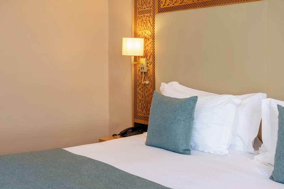 A neatly made bed with white and teal pillows, a bedside lamp, and a telephone on a wooden nightstand.