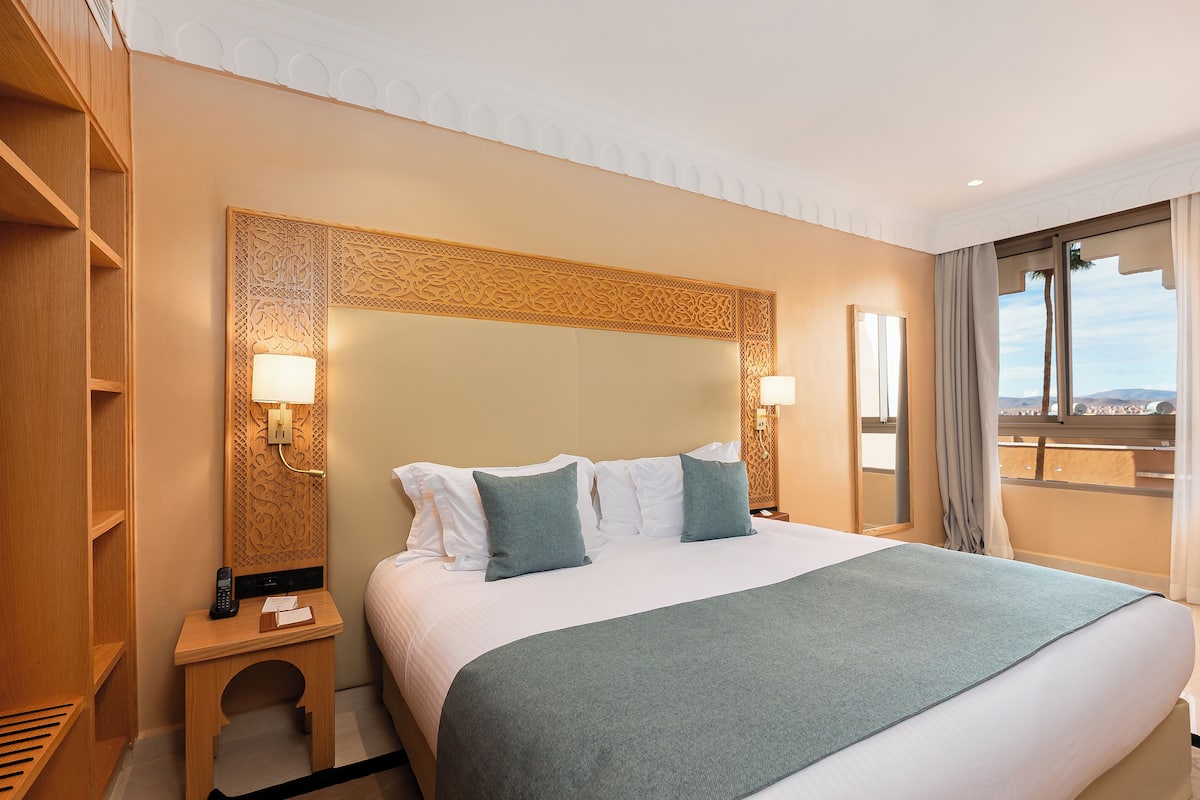 A hotel room with a large bed, two bedside lamps, a wooden nightstand, and a view of the outdoors.