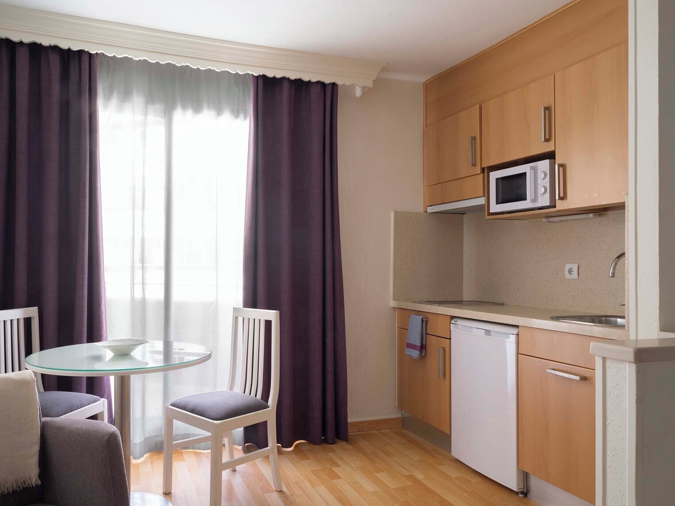 Cocina compacta con microondas, lavavajillas y armarios de madera. Hay una mesa redonda de vidrio con dos sillas y una ventana con cortinas moradas.