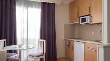 Cocina compacta con microondas, lavavajillas y armarios de madera. Hay una mesa redonda de vidrio con dos sillas y una ventana con cortinas moradas.
