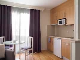 A compact kitchen with a microwave, dishwasher, and wooden cabinets. There is a round glass table with two chairs and a window with purple curtains.
