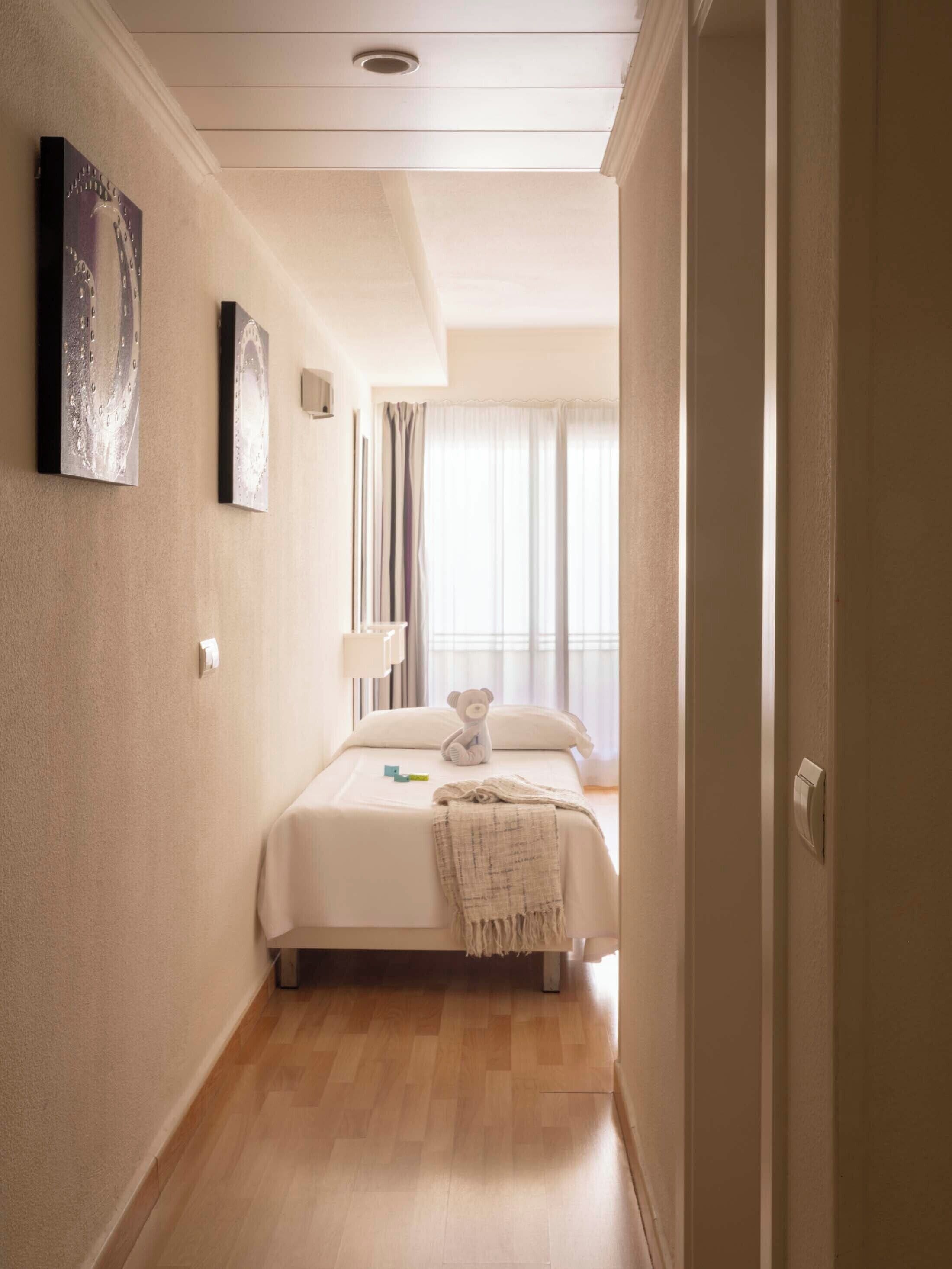 A hotel room with a bed, a bedside table, a lamp, a teddy bear, and a window with curtains.