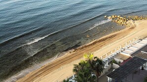 A beach with clear water, a sandy shore, and a building with a roofed area. - Ona Princesa Playa (Marbella)