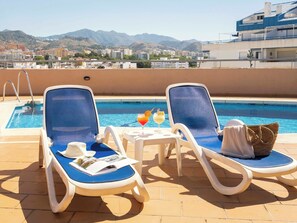 A poolside area with two lounge chairs, a small table with drinks, and a view of mountains in the background.