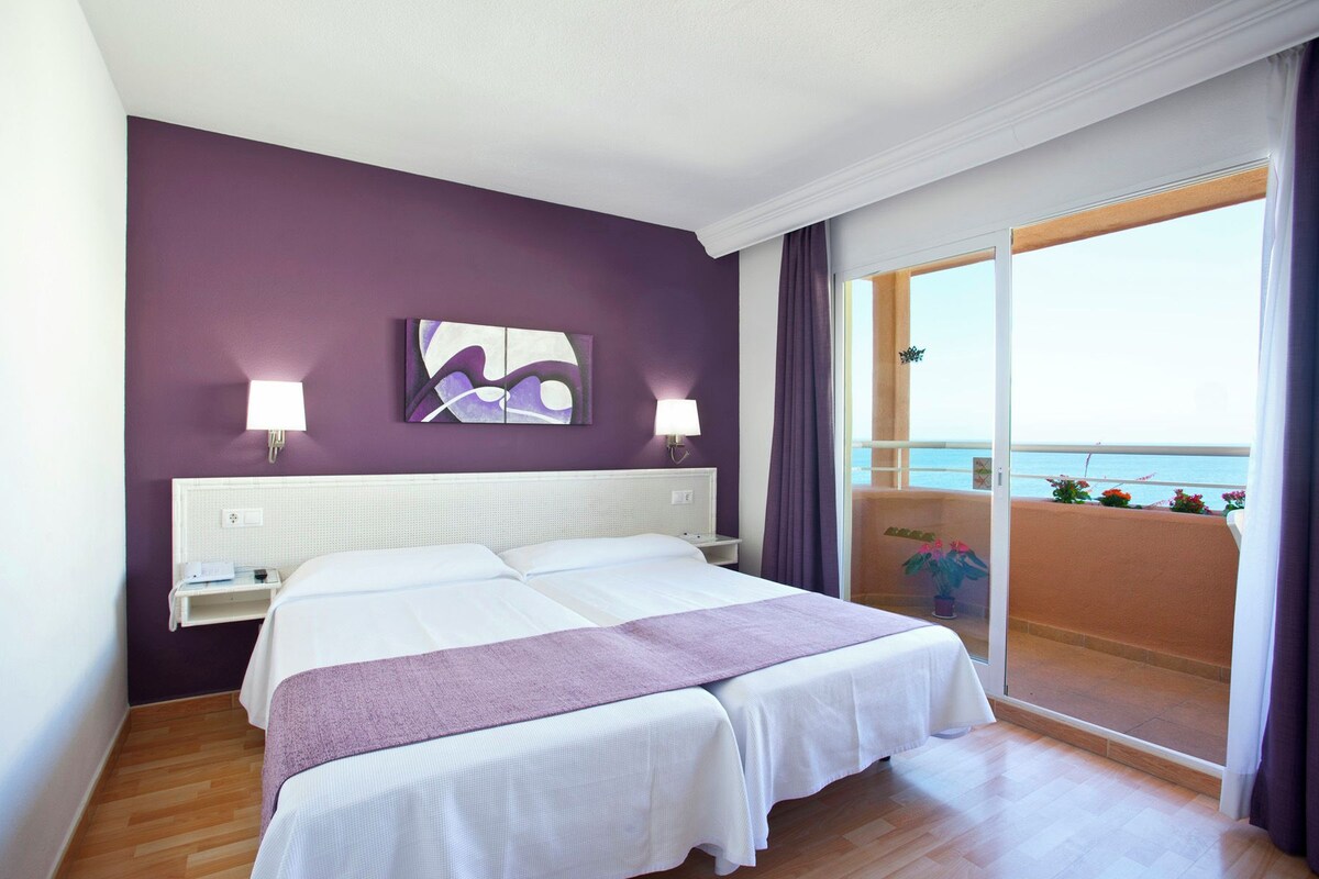 A hotel room with a large bed, a nightstand, a wall-mounted lamp, and a view of the sea through a balcony.