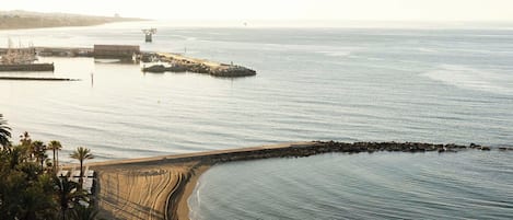 Vista costera con una playa de arena, un rompeolas y un puerto deportivo.