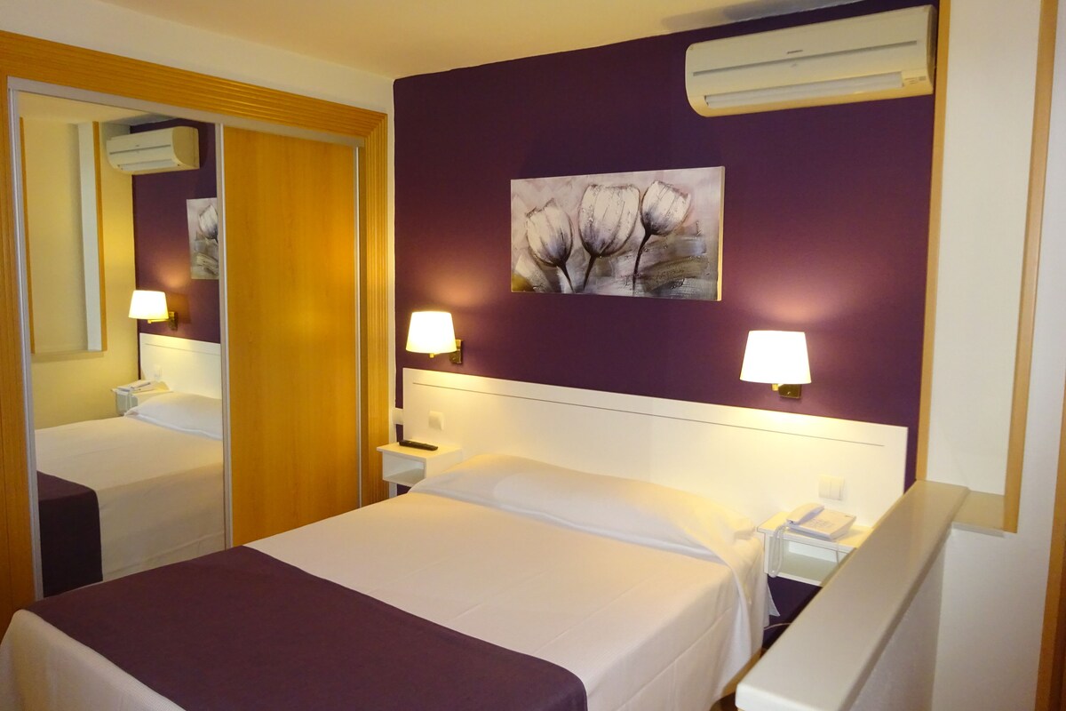 A hotel room with a bed, two bedside tables, a nightstand, a mirror, and an air conditioning unit on the wall.