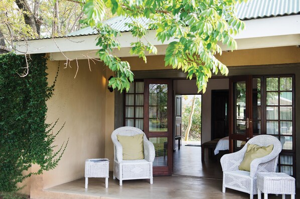 A covered outdoor seating area with wicker chairs and a small table, surrounded by greenery and a building entrance.