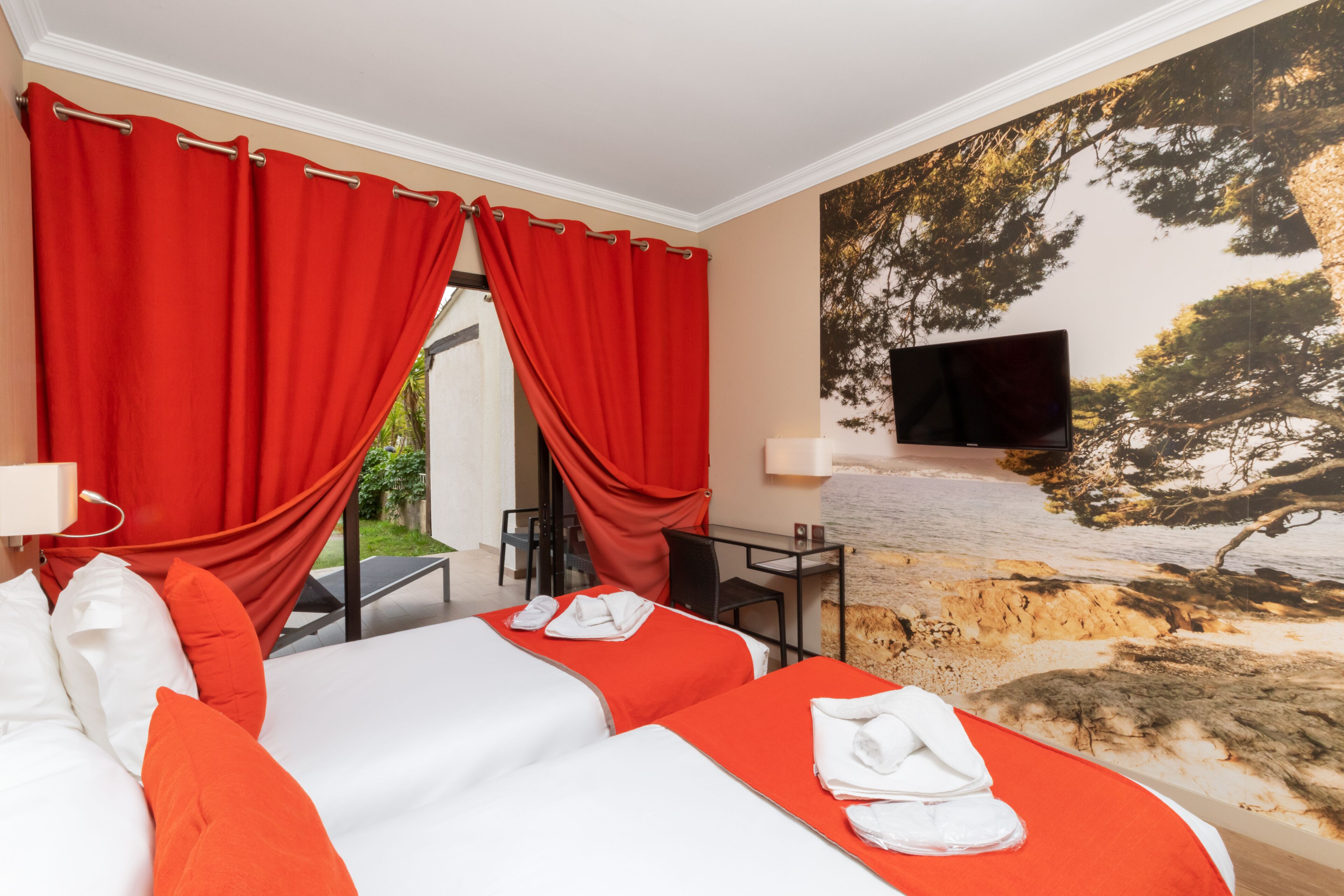 Habitación de hotel con dos camas, cortinas rojas, un televisor, un escritorio y un mural con árboles.