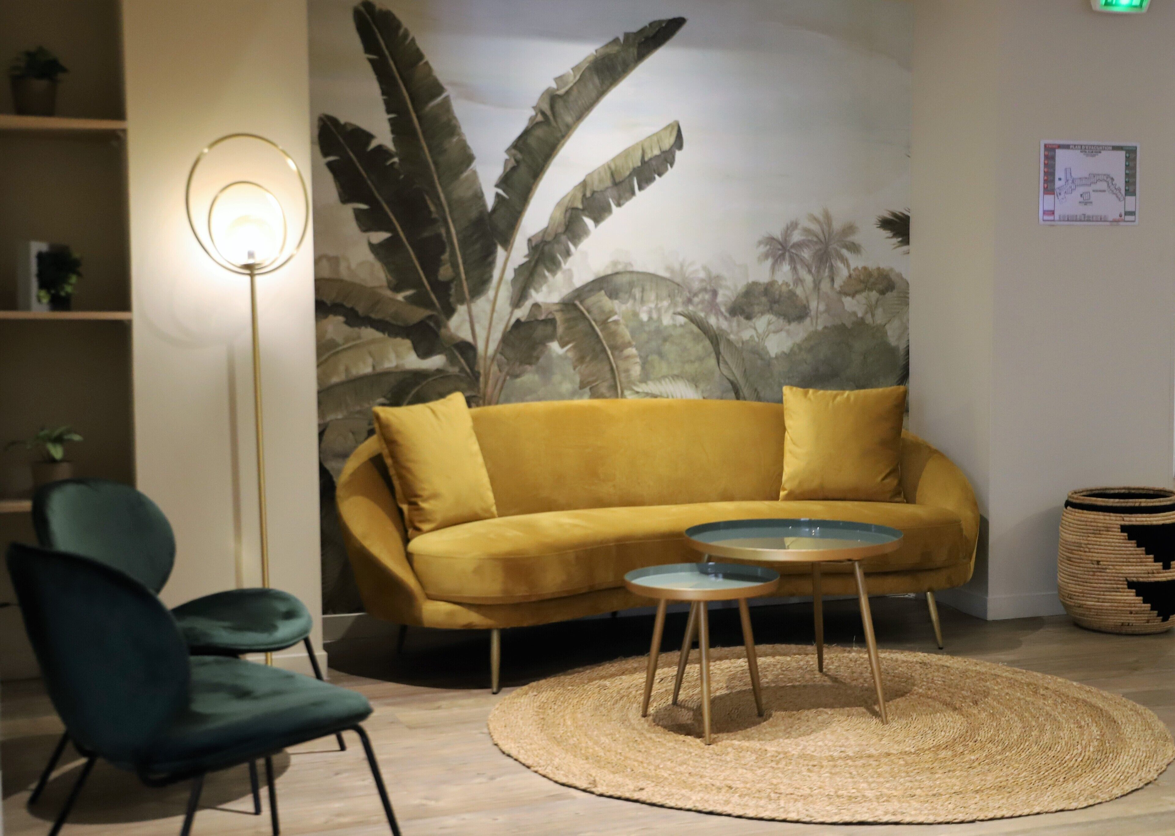 Une pièce avec un canapé jaune moutarde, un fauteuil vert, une petite table ronde, un lampadaire et une fresque murale représentant des feuillages.