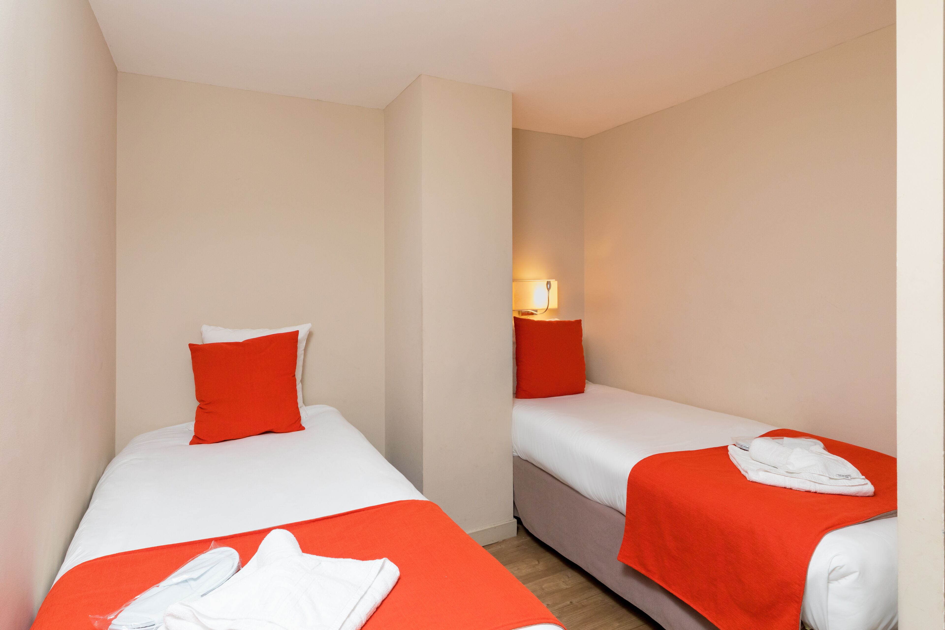 Une chambre d’hôtel avec deux lits simples, chacun recouvert d’une couverture rouge et agrémenté d’oreillers blancs. Une table de chevet avec une lampe se trouve entre les deux lits.