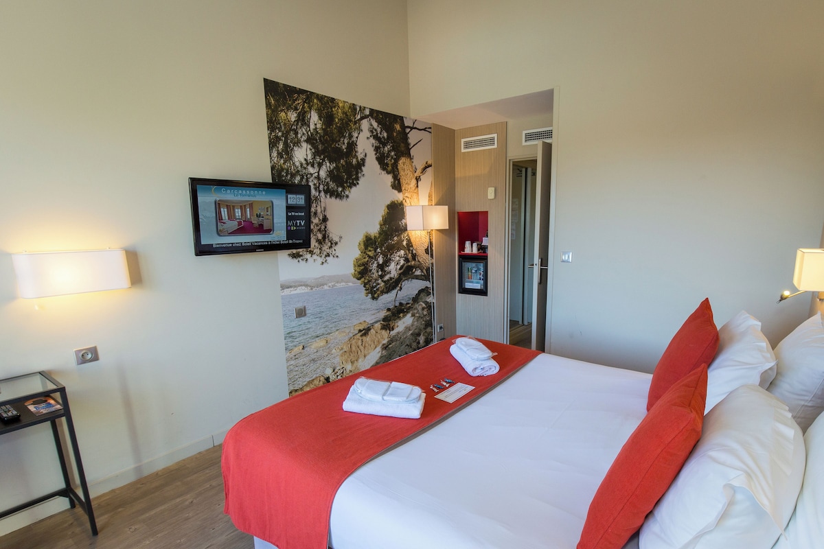 A hotel room with a bed, a nightstand, a wall-mounted TV, and a painting of a beach scene.