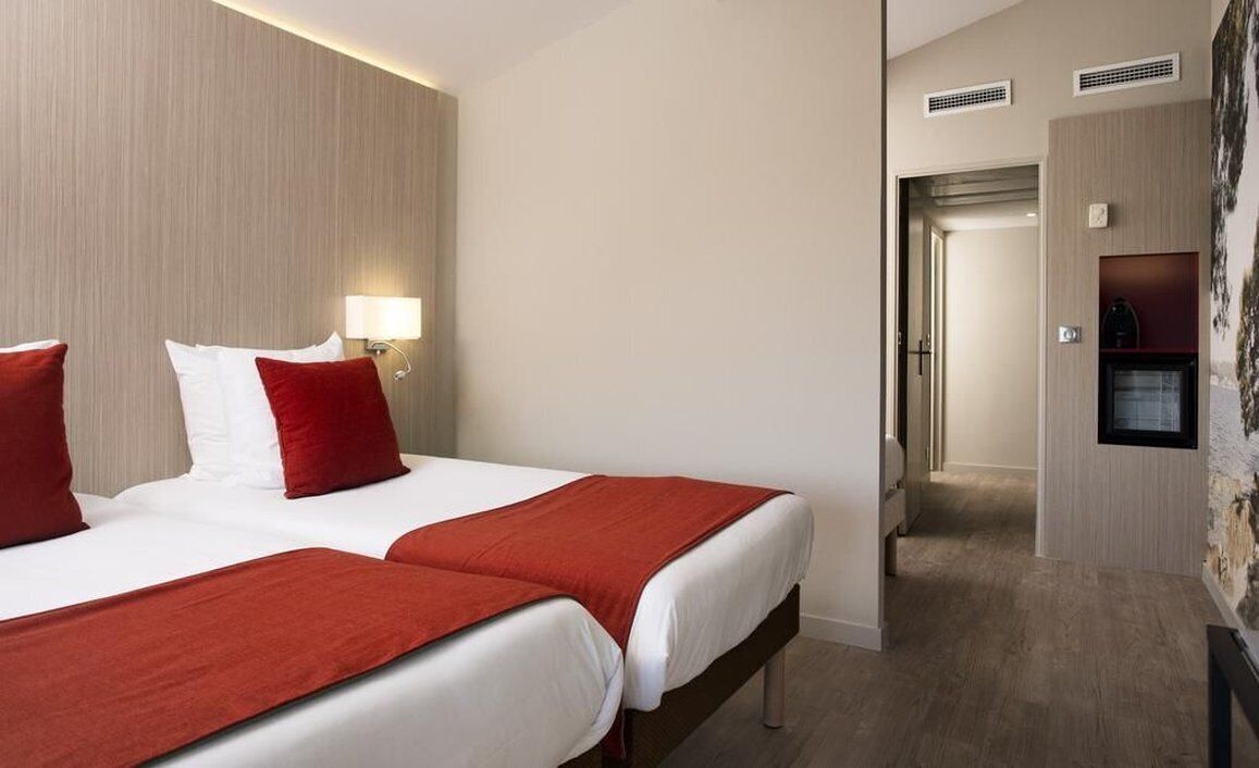 A hotel room with two beds, a red and white bedspread, a wall-mounted lamp, and a wooden headboard.