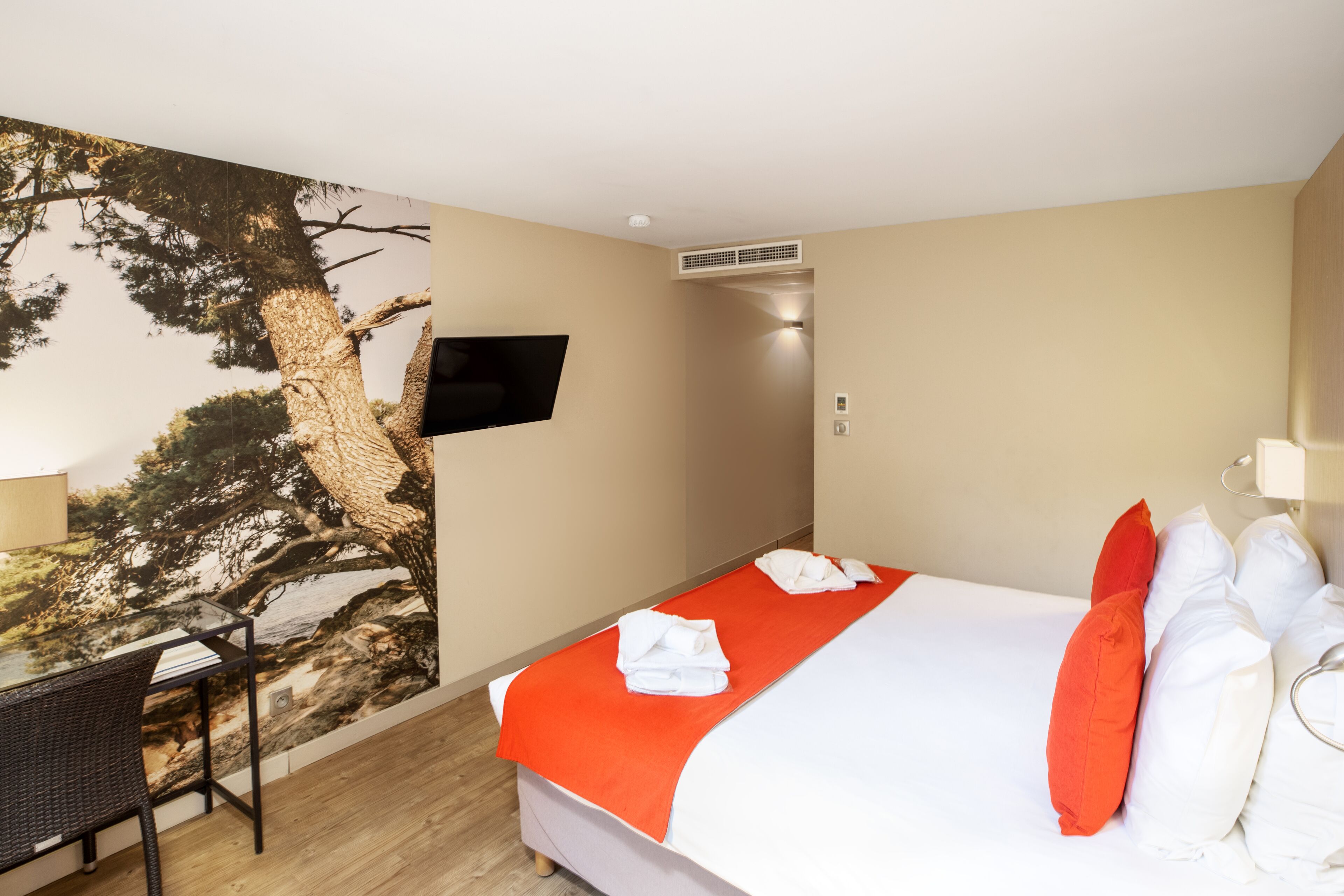 A hotel room with a bed, a desk, a chair, a TV, and a mural of a tree.