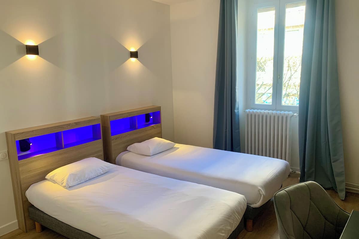 A room with two beds, a chair, a window with curtains, and a wall-mounted light fixture.
