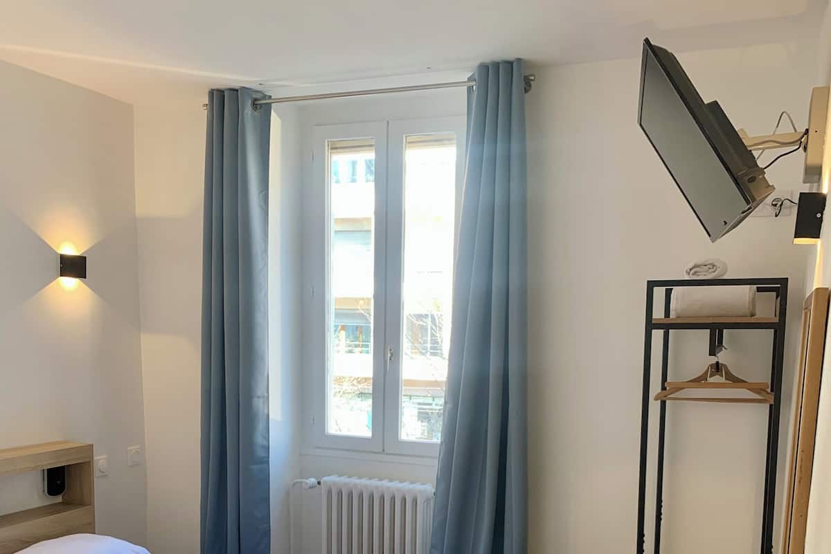 A hotel room with a bed, a desk, a chair, a hanging light, and a window with curtains.