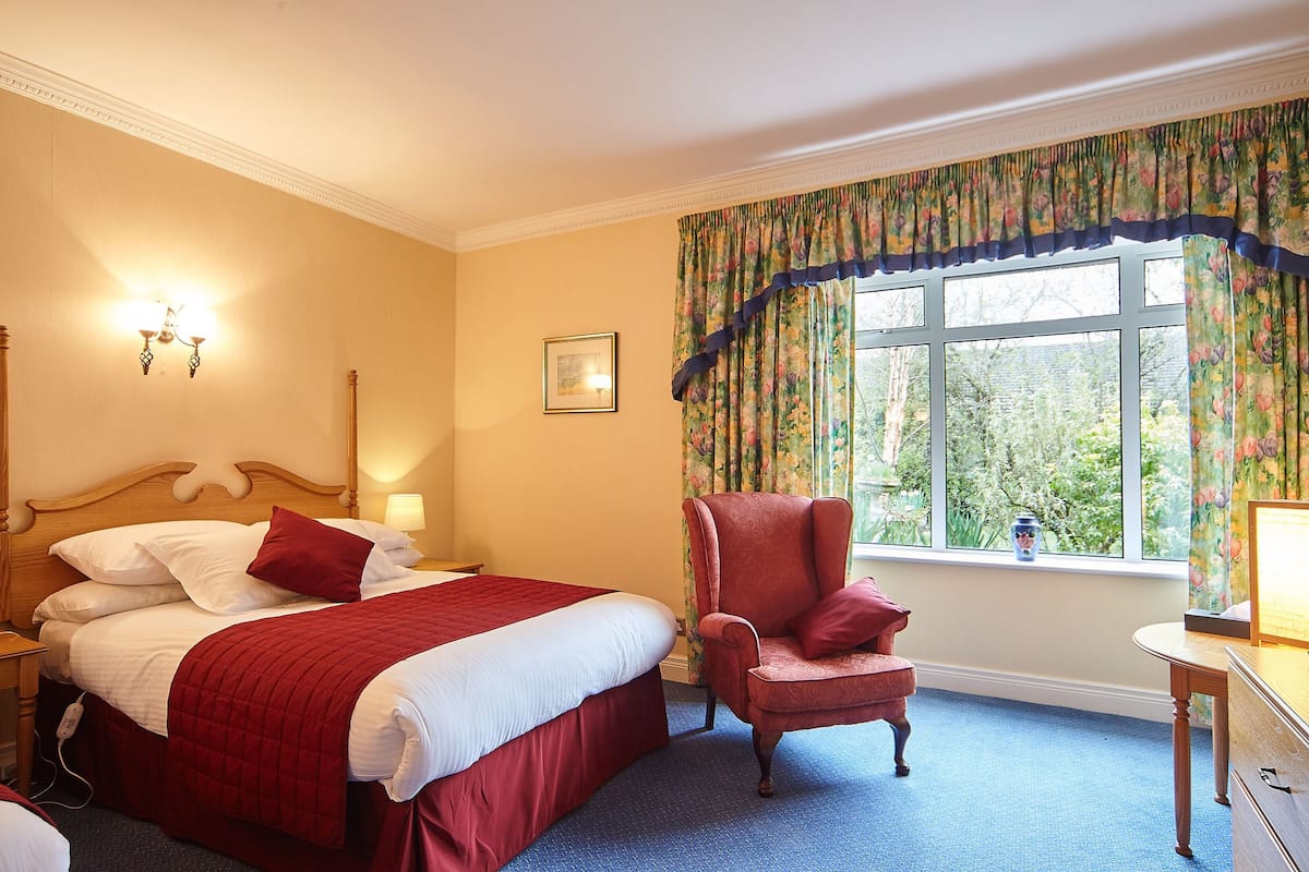Une chambre d’hôtel avec un grand lit, une chaise, un bureau et une fenêtre agrémentée de rideaux à motifs floraux.