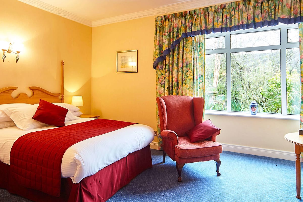 Une chambre d’hôtel avec un lit, une chaise, un bureau et une fenêtre agrémentée de rideaux à motifs floraux.