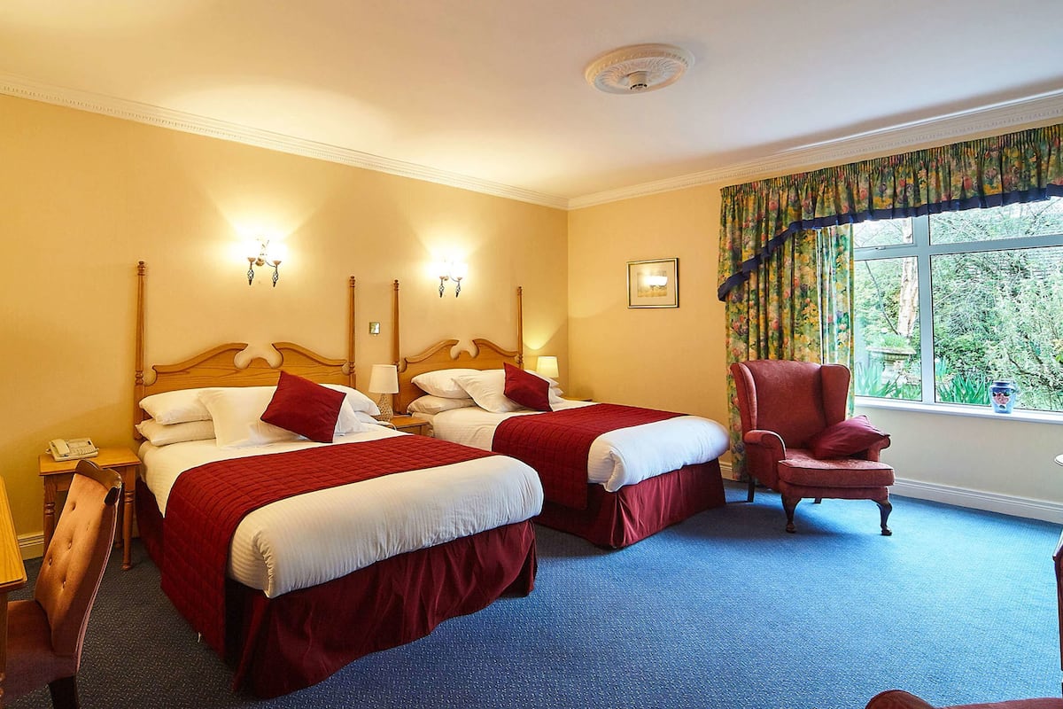 Une chambre d’hôtel avec deux lits, un bureau, une chaise et une fenêtre donnant sur un espace verdoyant.