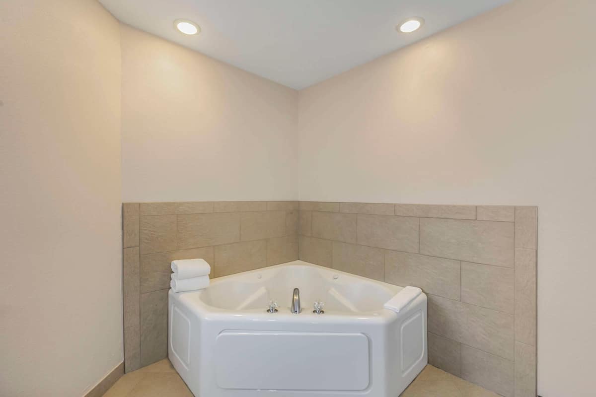 A corner bathroom with a white bathtub, beige tiled walls, and recessed ceiling lights.