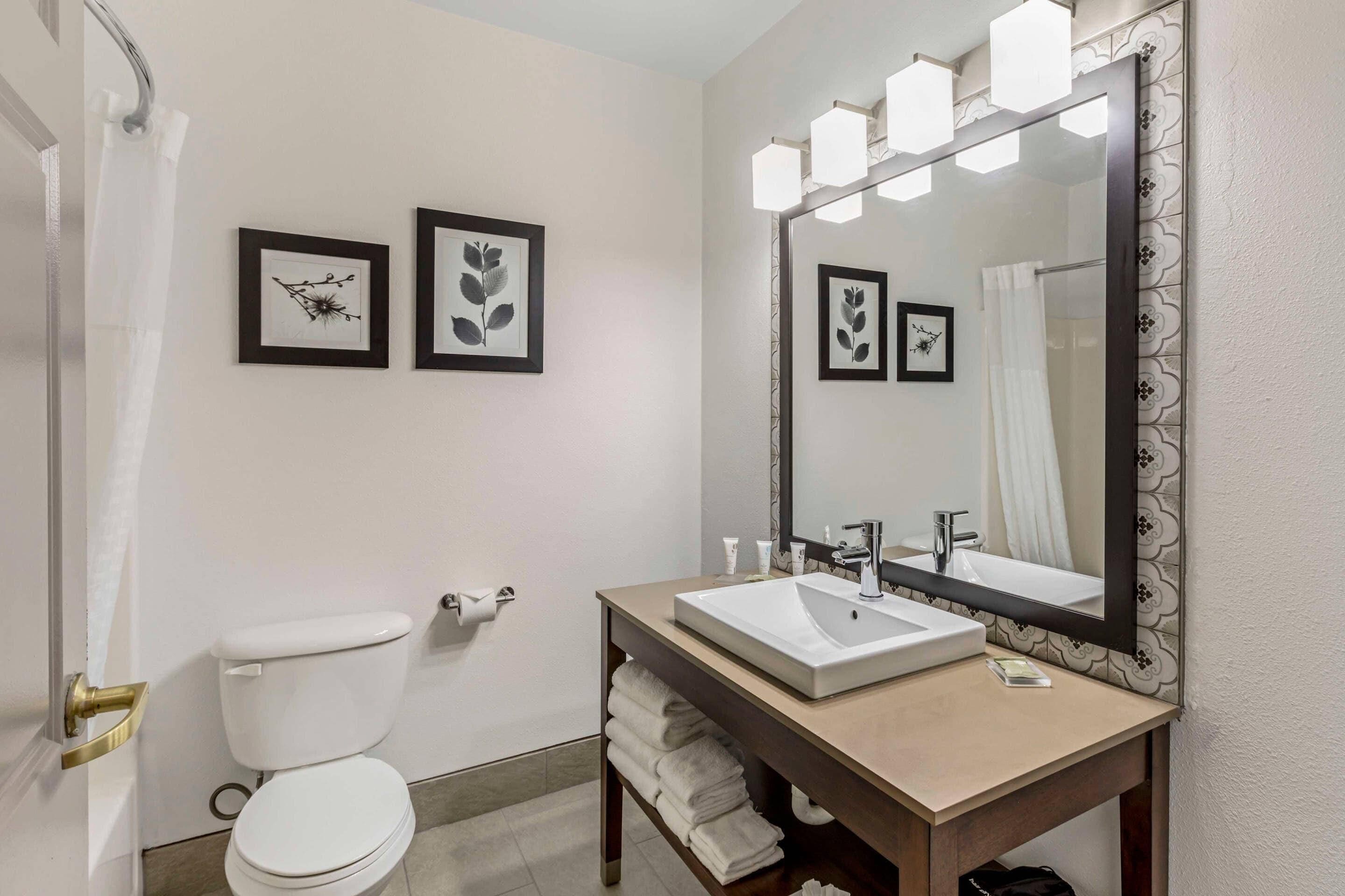 A modern bathroom with a toilet, sink, and large mirror. There are framed pictures on the wall and a towel rack.