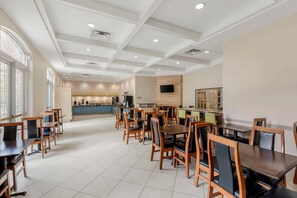 A spacious dining area with multiple tables and chairs, a kitchen in the background, and large windows allowing natural light.