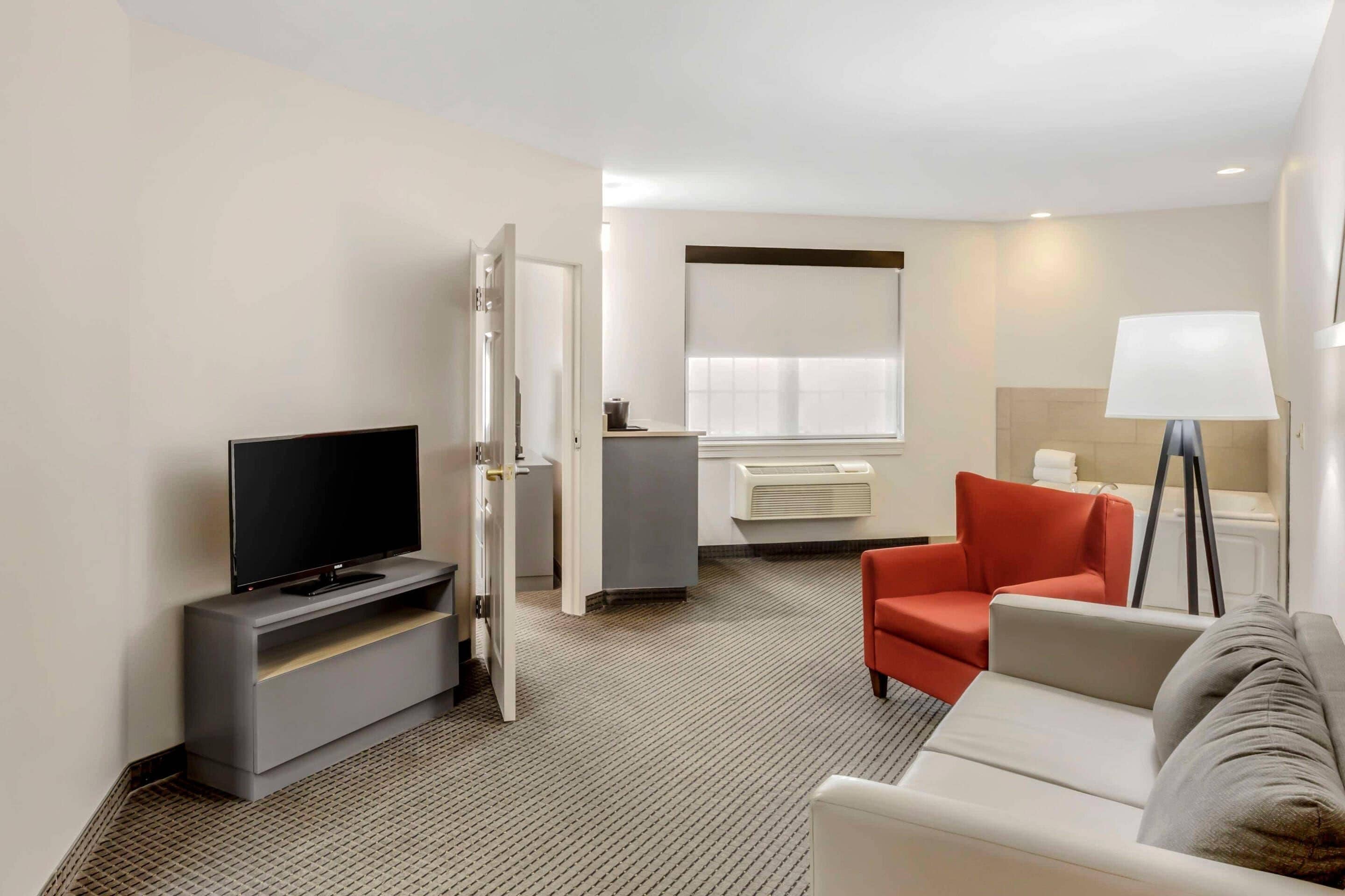 A hotel room with a TV, a sofa, a red chair, a lamp, and a window with blinds.