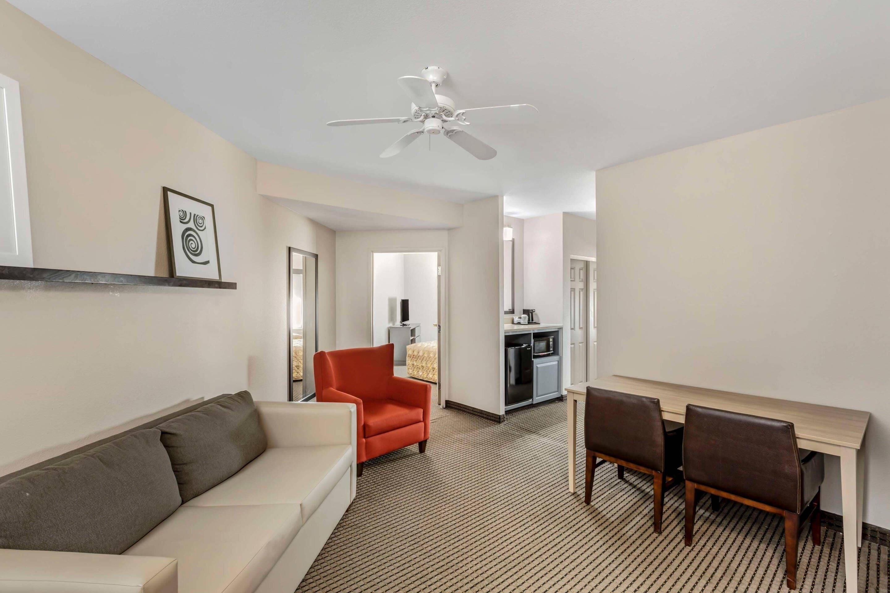A hotel room with a sofa, a dining table with chairs, a ceiling fan, and a kitchenette in the background.