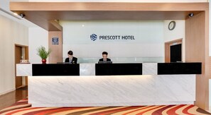 Un comptoir d’accueil à l’hôtel Prescott, avec deux employés, une horloge et une enseigne indiquant le nom de l’hôtel.