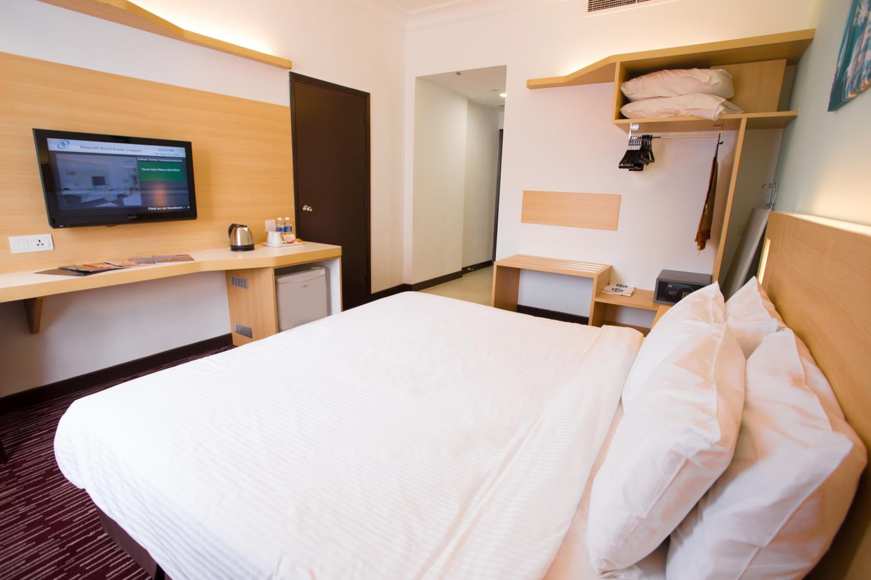 Une chambre d’hôtel avec un lit, un bureau avec un ordinateur, une télévision et une étagère avec des serviettes.