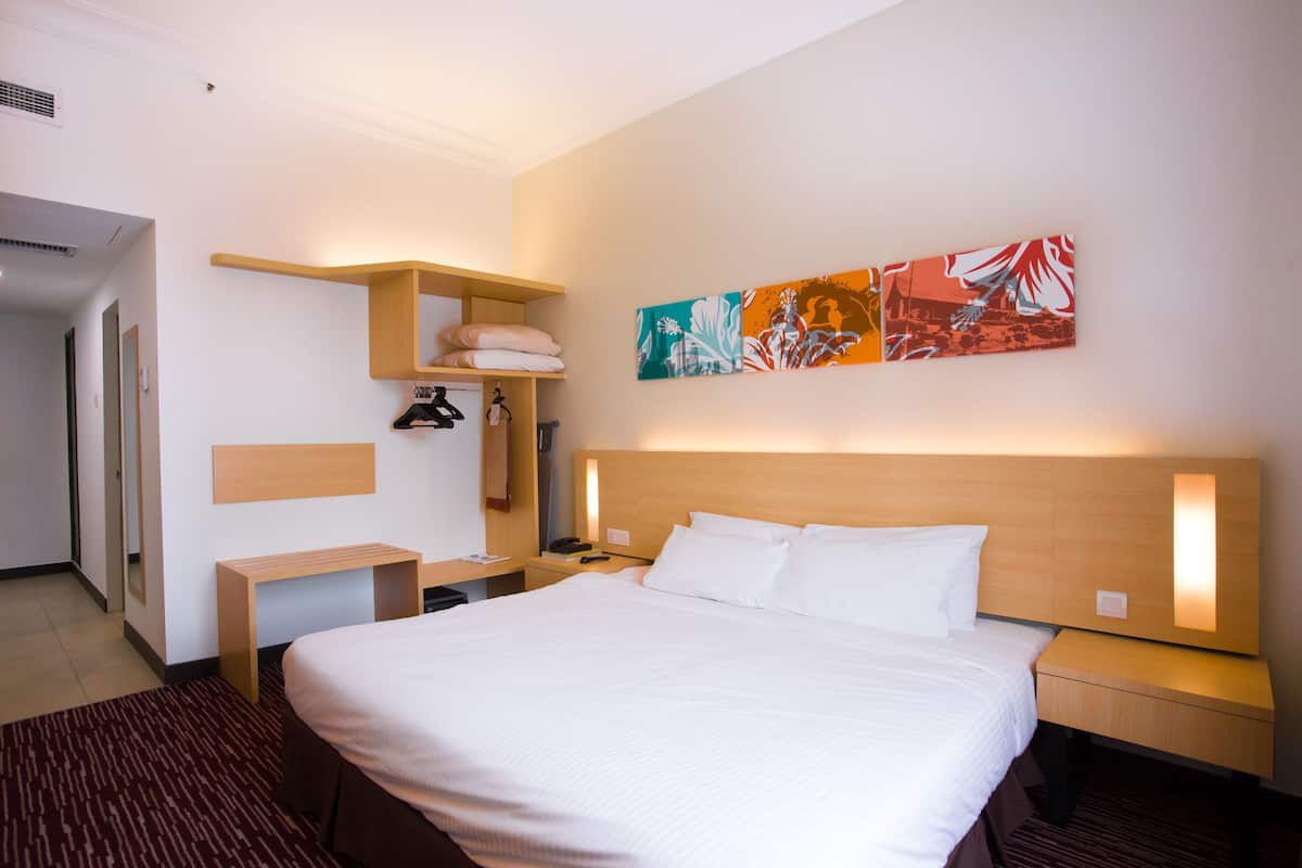 A hotel room with a large bed, bedside tables, a shelf with towels, and artwork on the wall.
