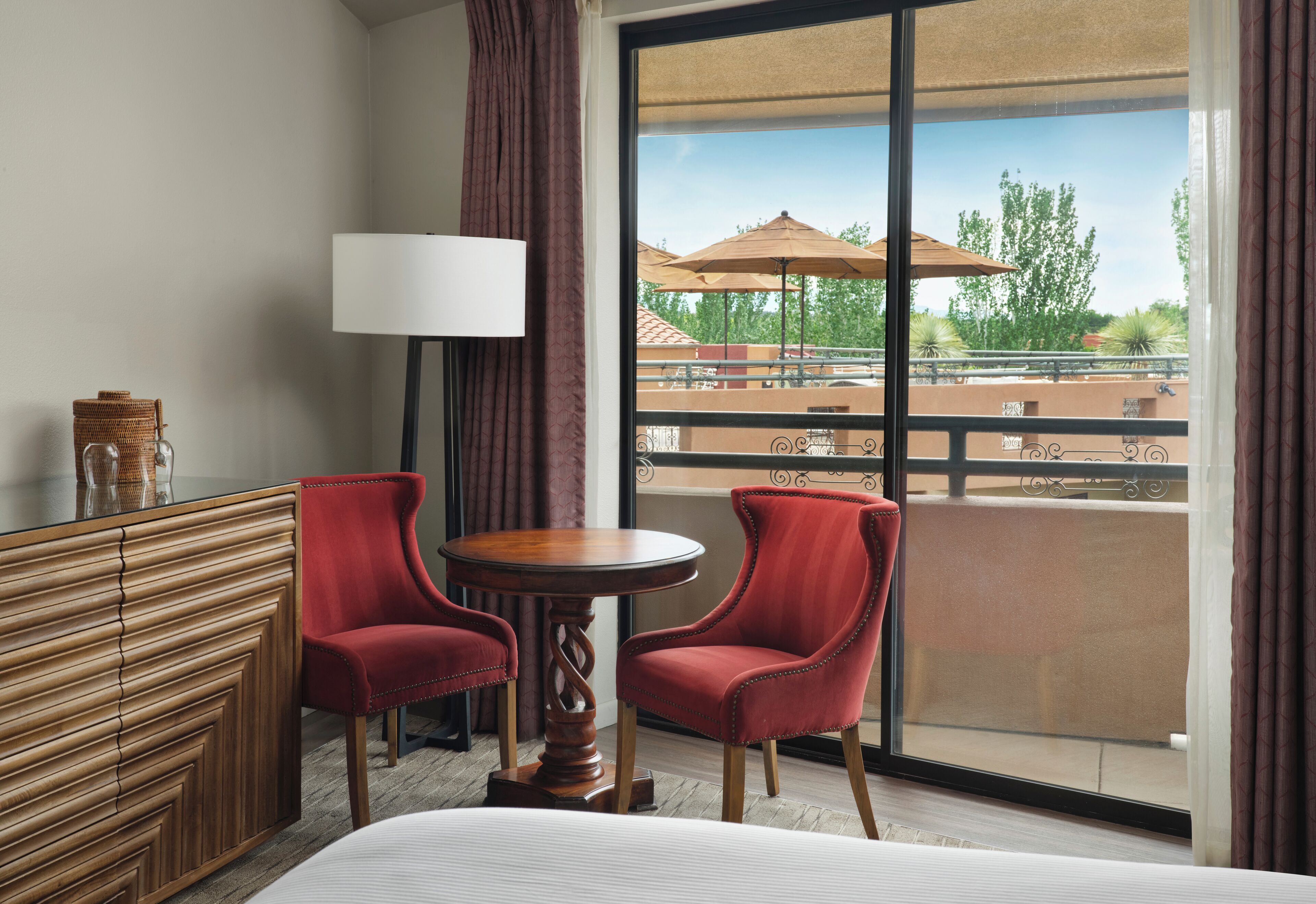 A hotel room with a bed, two red chairs, a small round table, a lamp, and a large window with a view of outdoor seating and trees.
