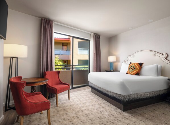 Habitación de hotel con una cama grande, dos sillas rojas, una mesita pequeña, un balcón con vistas a edificios y una ventana con cortinas.