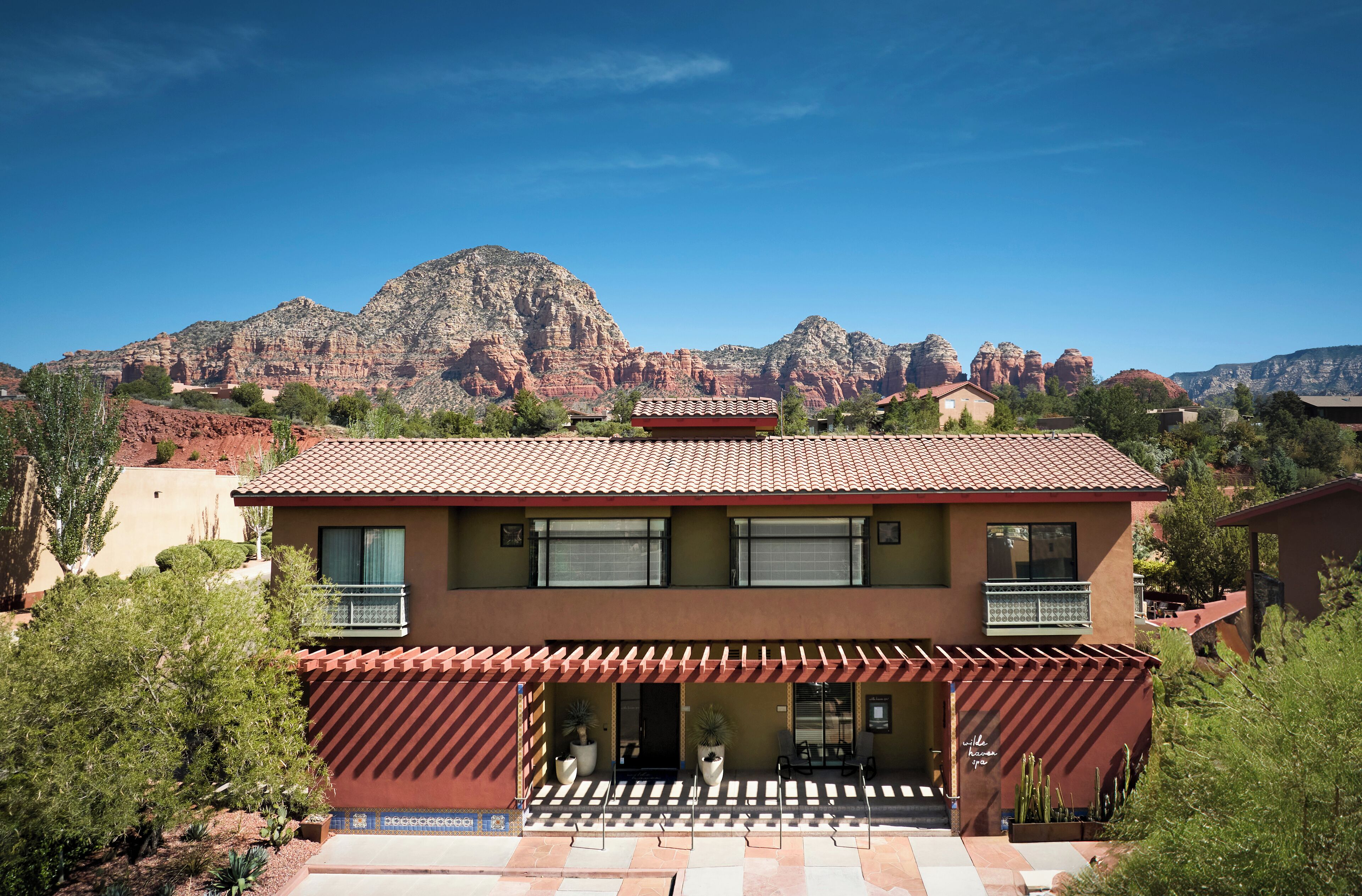 A two-story building with a red-tiled roof, a covered patio with outdoor seating, and a view of red rock formations in the background.