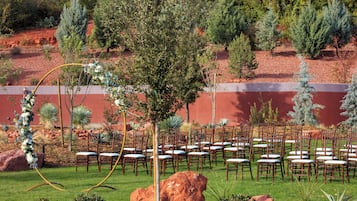 Un espace extérieur aménagé, avec des rangées de chaises et de tables en bois, entouré de jardins paysagers et d’arbres.