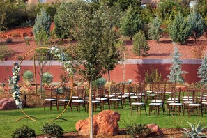 A formal outdoor seating area with rows of wooden chairs and tables, surrounded by landscaped gardens and trees.