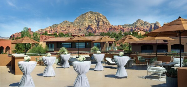 Un patio en la azotea con mesas redondas, sillas y sombrillas, con vistas a un paisaje montañoso.