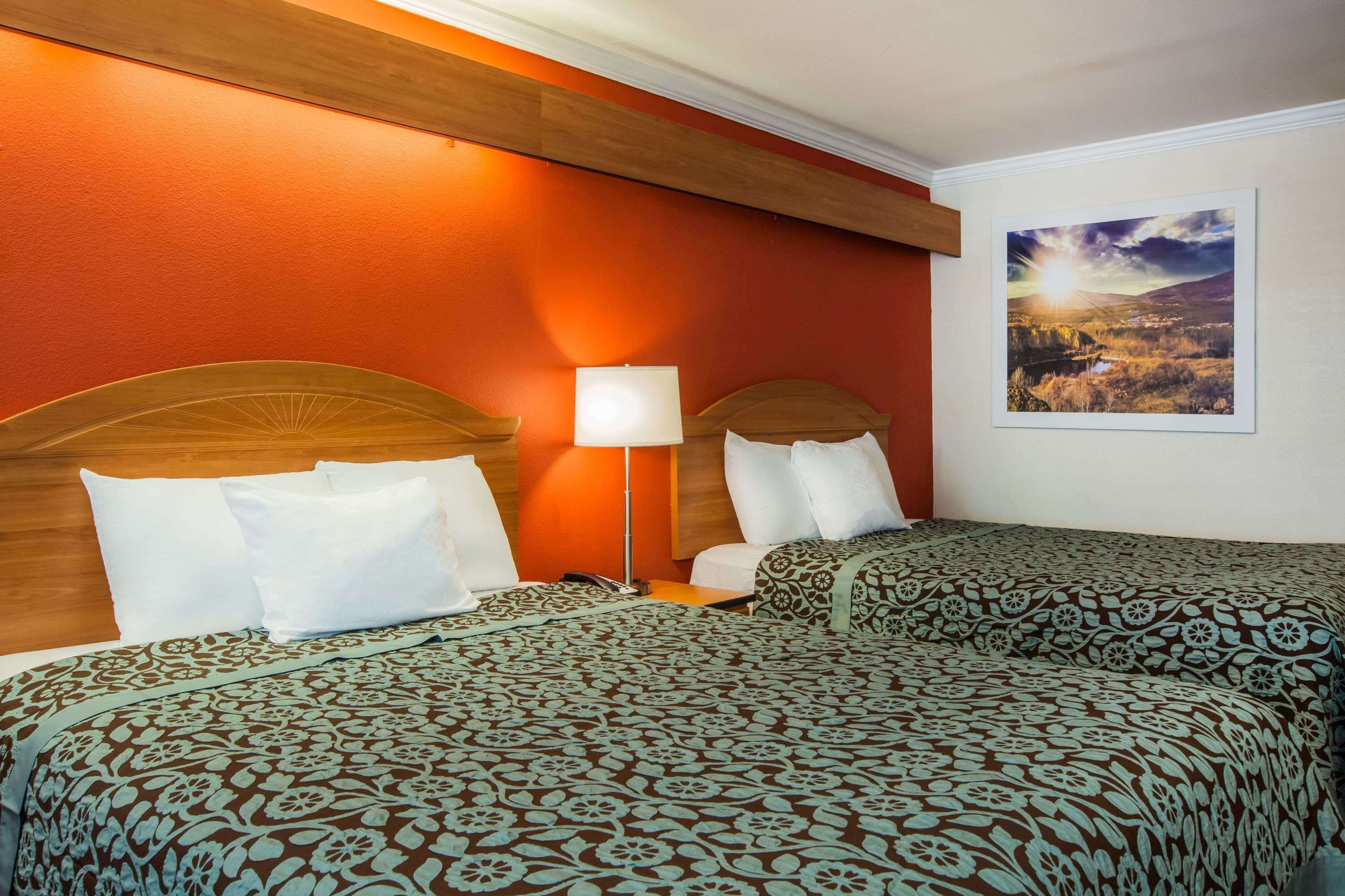 A hotel room with two beds, a wooden headboard, a nightstand, a lamp, and a framed landscape painting on the wall.