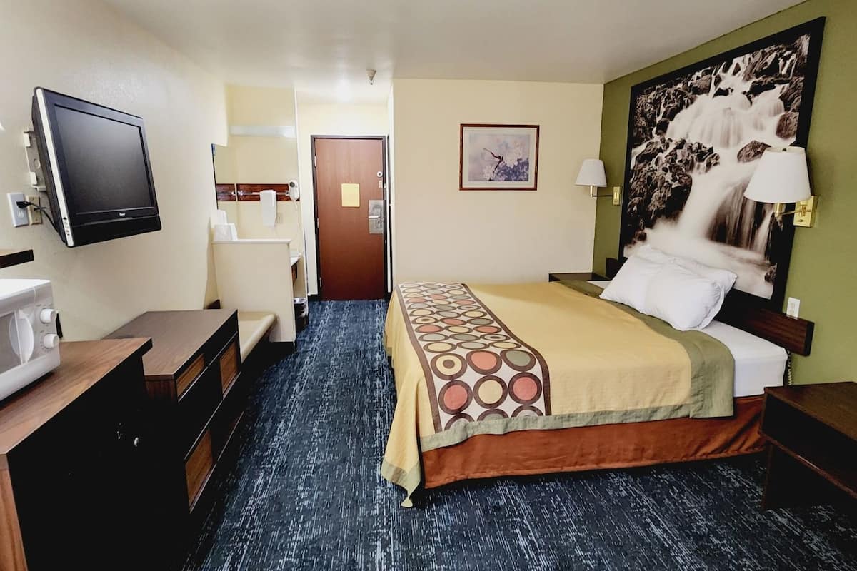 A hotel room with a bed, a TV, a microwave, a desk, a chair, and a bathroom visible in the background.