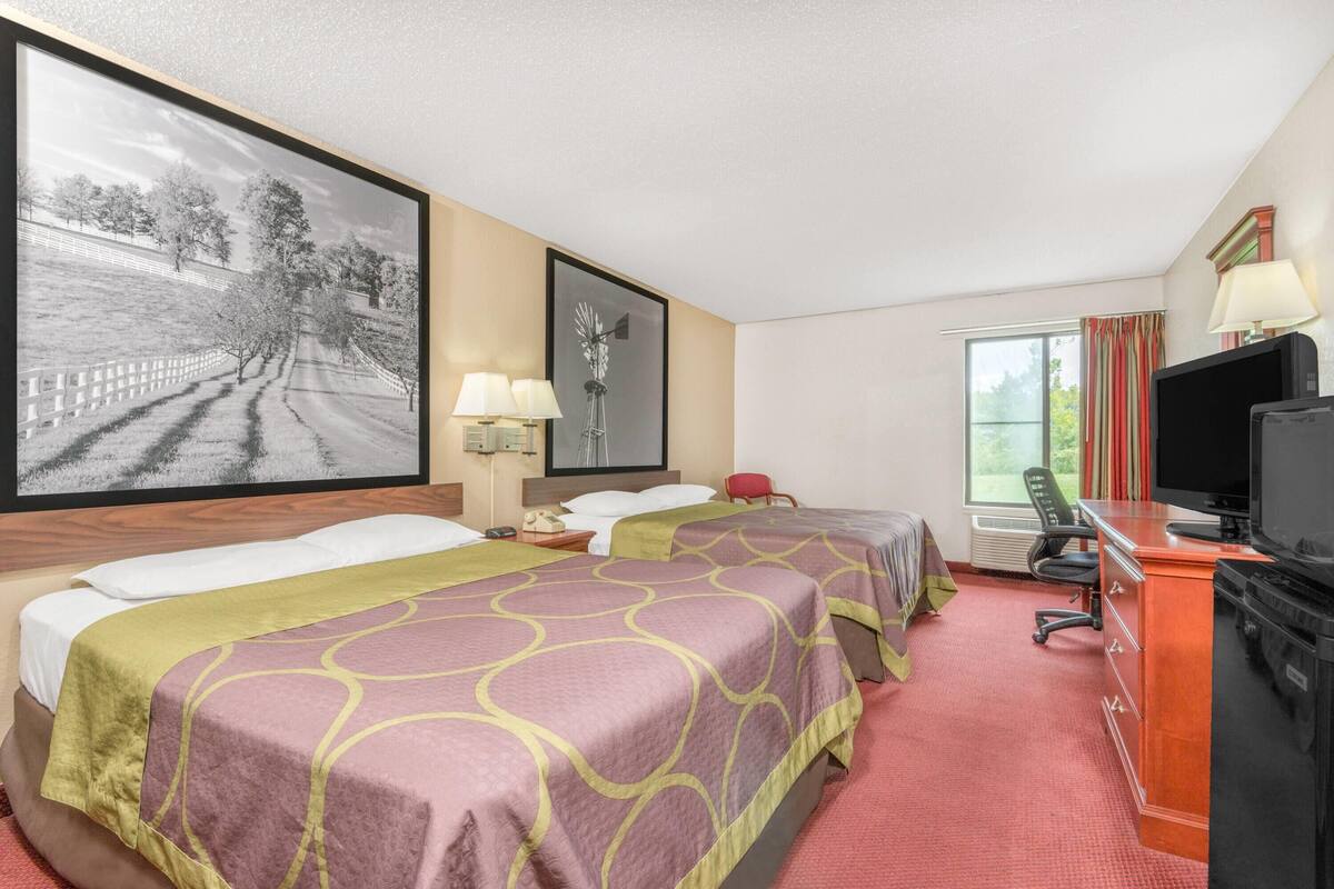 A hotel room with two beds, a desk with a television, a large framed picture, and a window with curtains.