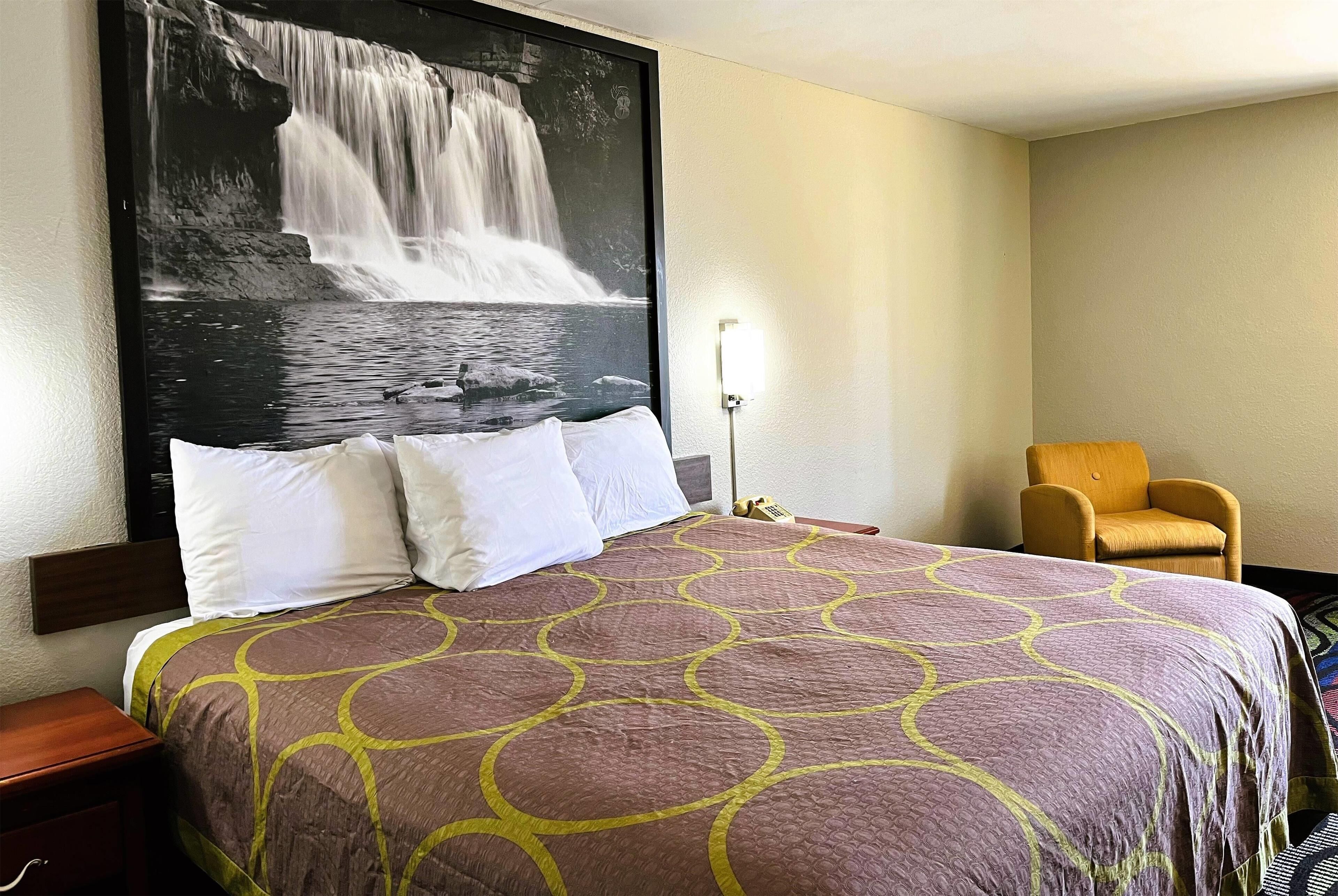 A hotel room with a large bed, a chair, a nightstand, and a wall-mounted lamp.