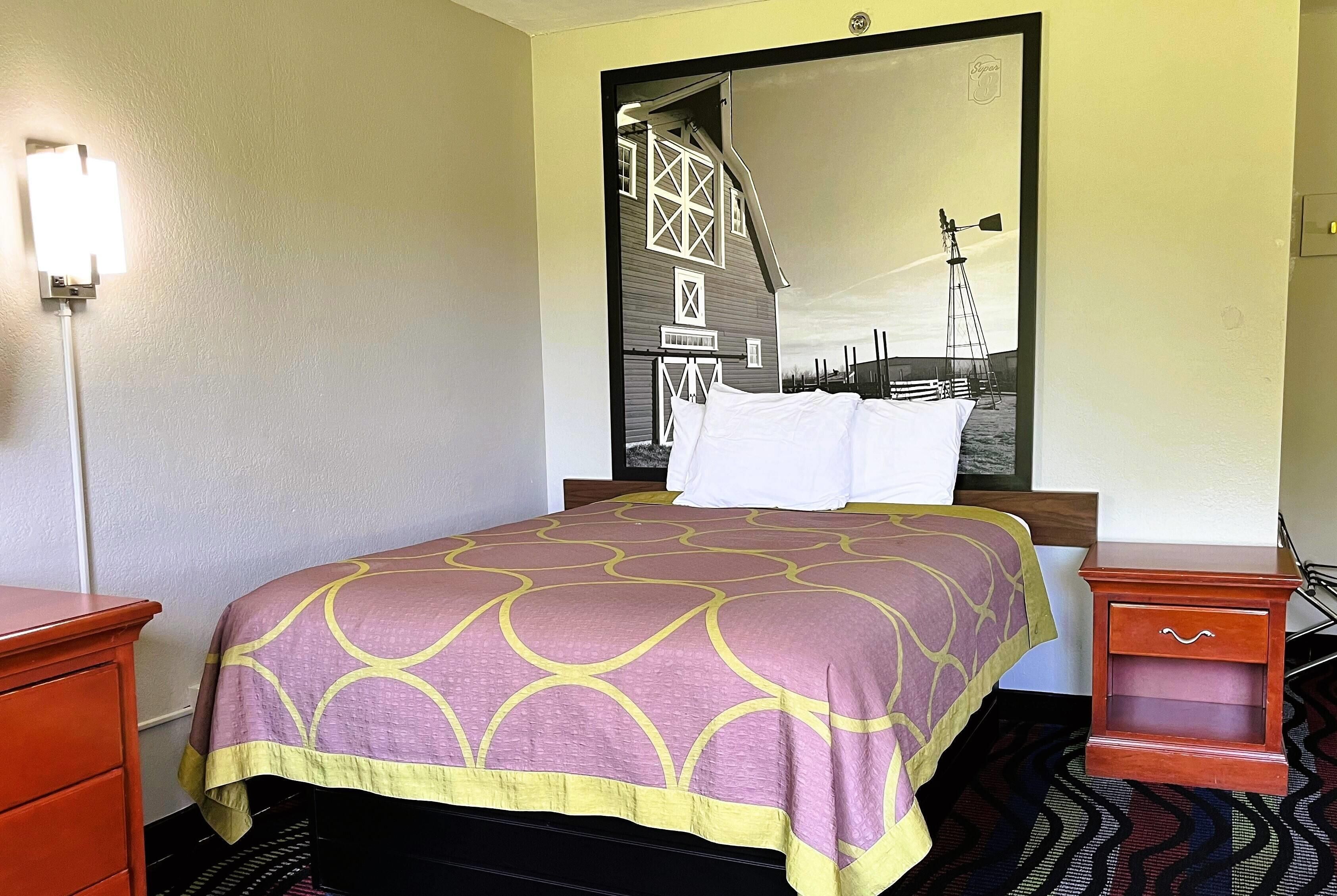 A hotel room with a bed, a nightstand, a dresser, and a large mirror reflecting a barn and windmill.