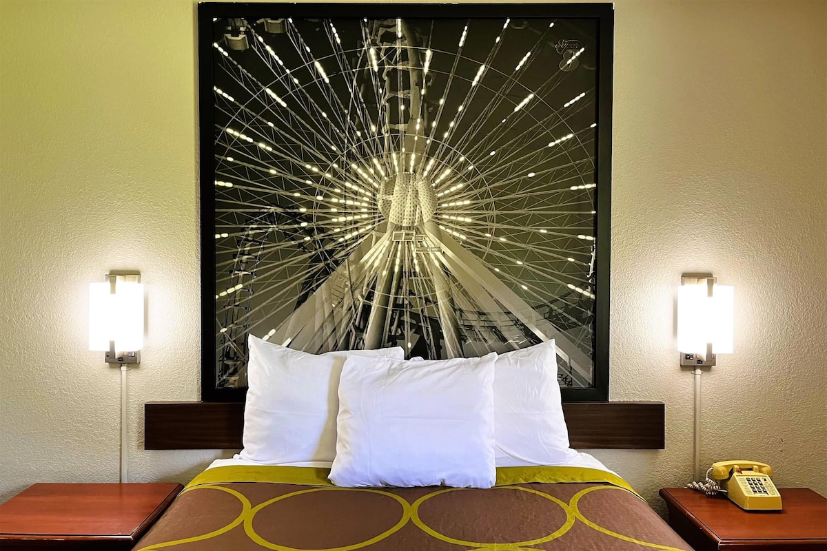 A hotel room with a bed, white pillows, a nightstand with a telephone, and a wall-mounted light fixture.