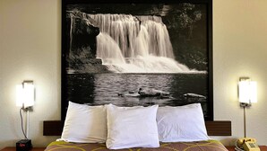 Habitación de hotel con cama, almohadas blancas, mesita de noche con un reloj, teléfono y una lámpara fijada a la pared.