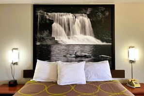 Ein Hotelzimmer mit einem Bett, weißen Kissen, einem Nachttisch mit einer Uhr, einem Telefon und einer Wandleuchte.