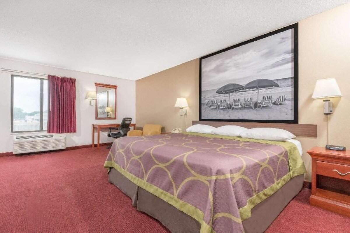 A hotel room with a bed, a desk, a chair, a window with curtains, a wall-mounted lamp, and a framed picture of a beach scene.