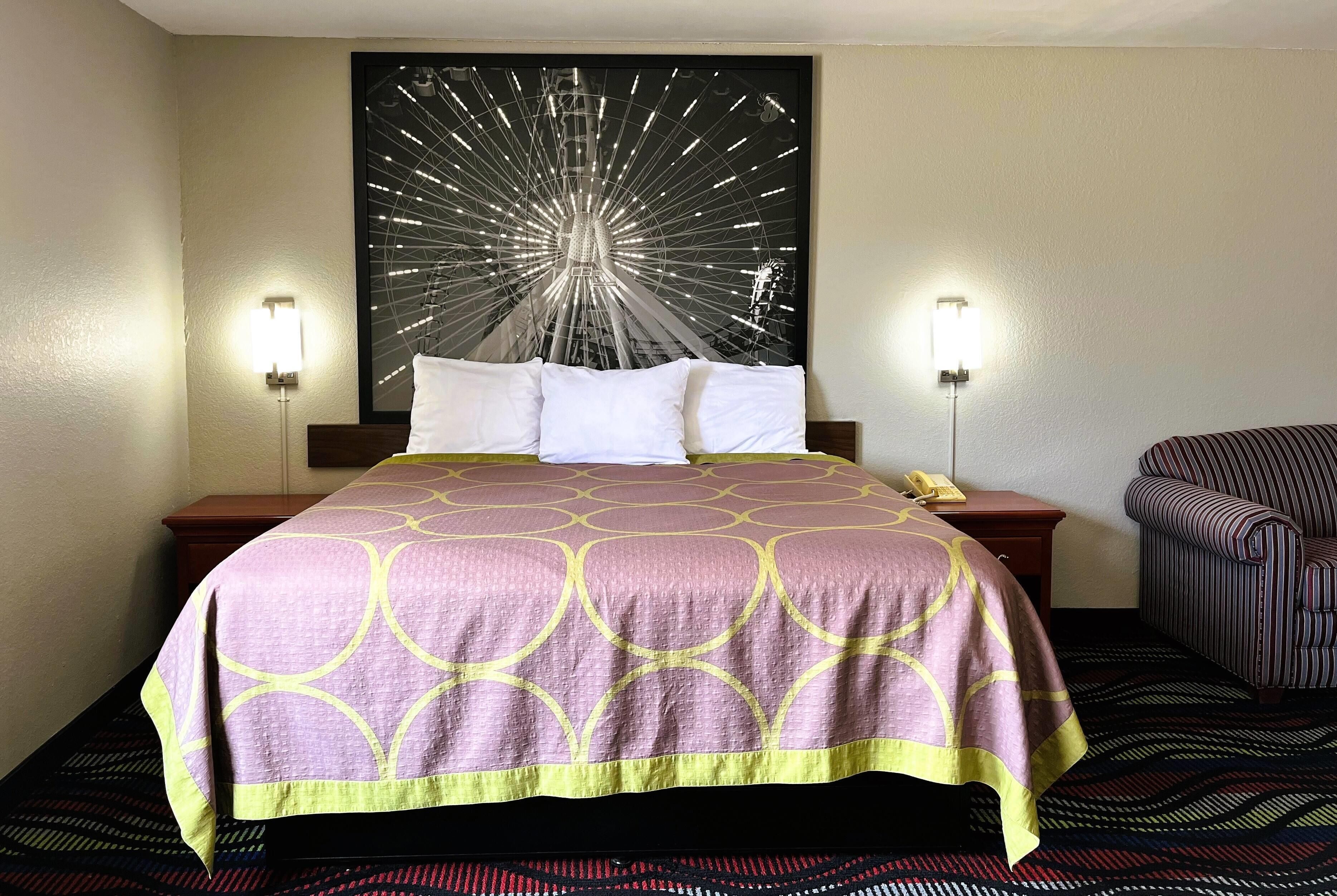 A hotel room with a bed, a headboard with a light fixture, a chair, and a wall-mounted artwork.