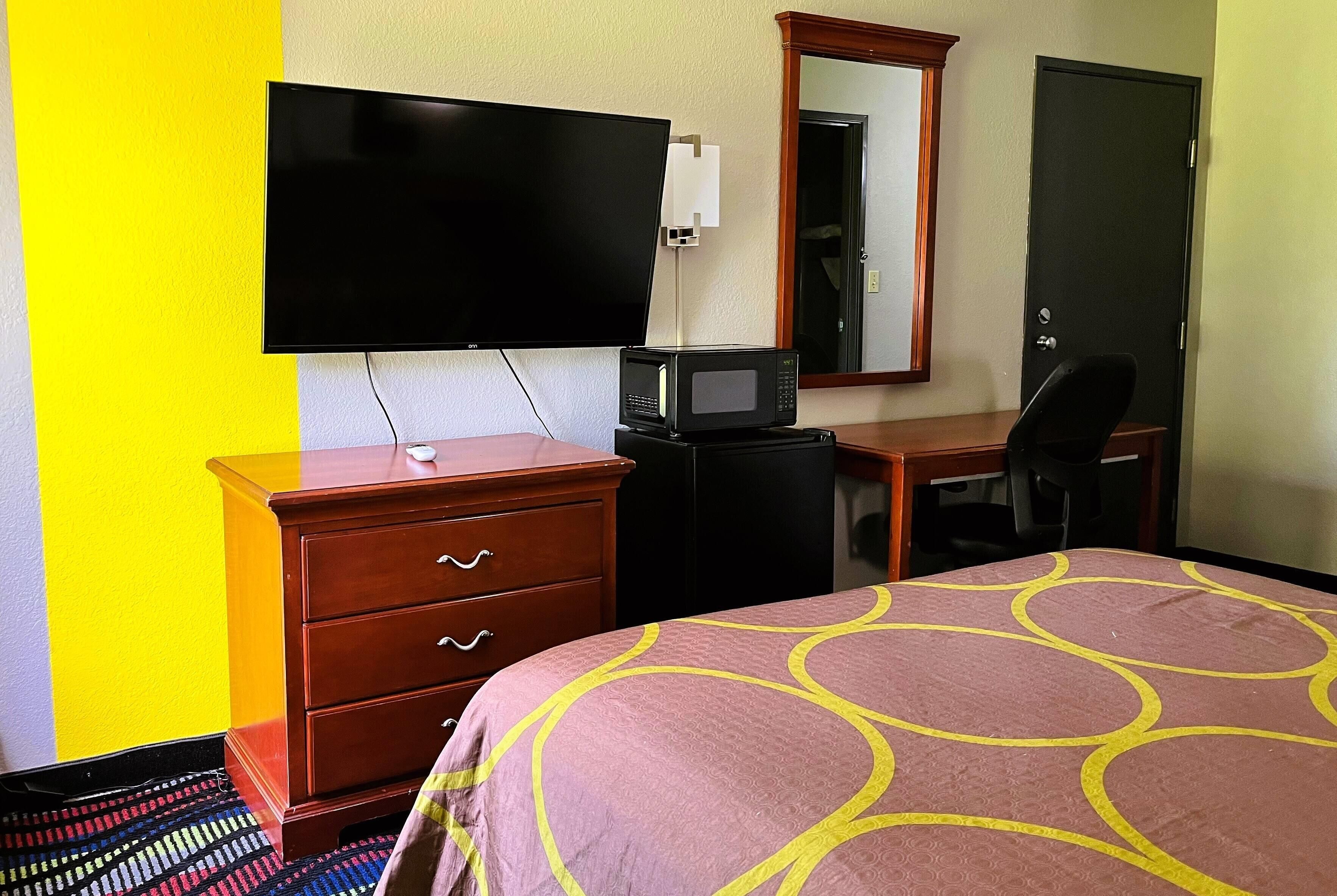 A hotel room with a bed, a desk, a television, a microwave, a dresser, and a door.