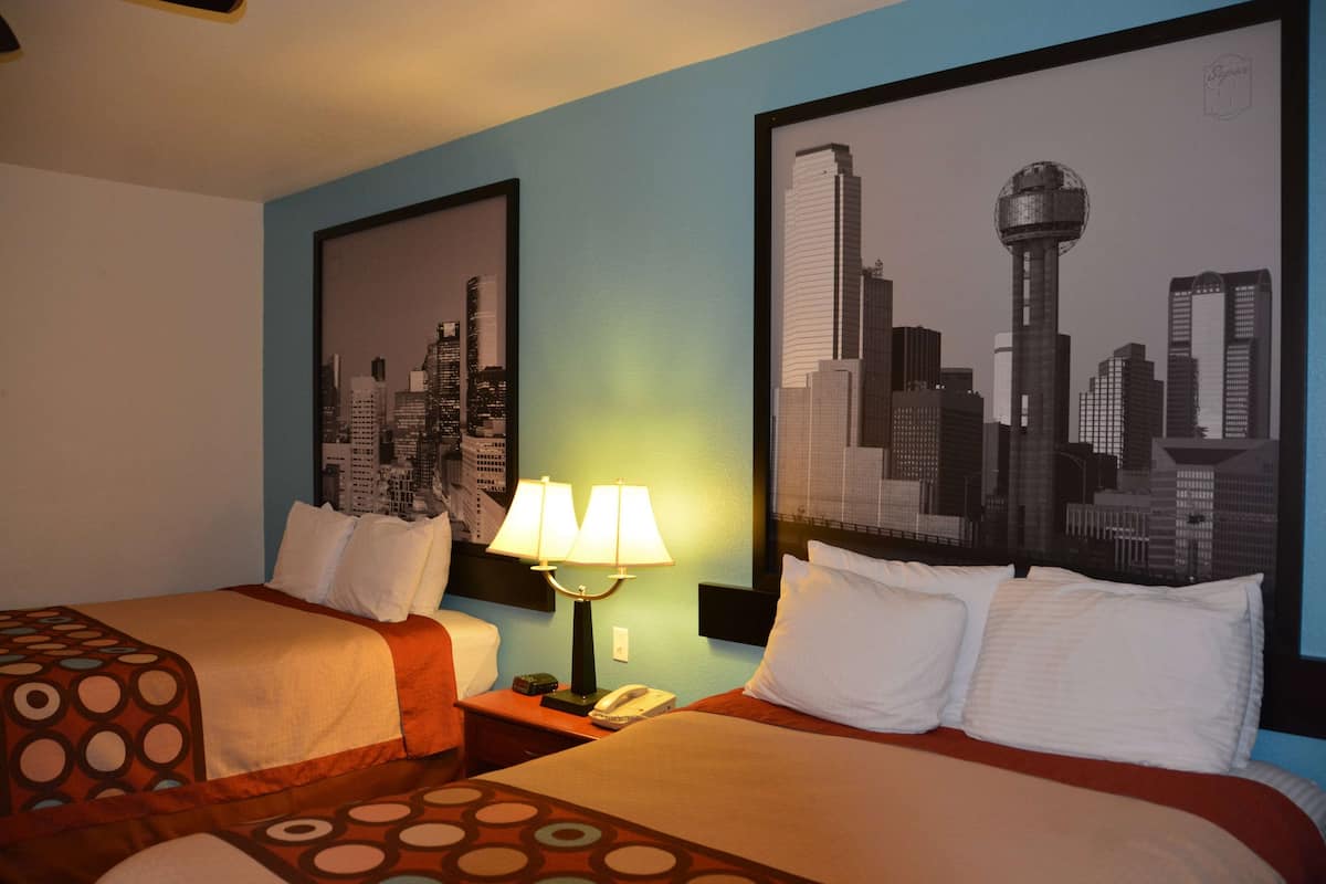Habitación de hotel con dos camas, una mesita de noche con lámpara y dos cuadros enmarcados con vistas a la ciudad.